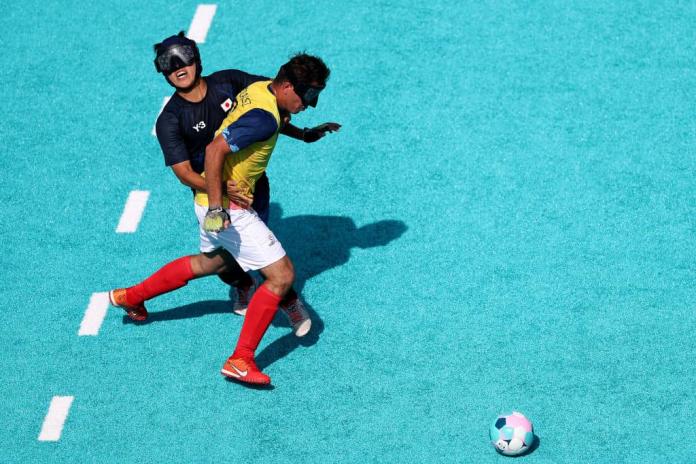Two footballers wearing blindfolds, one in yellow, one in blue, compete for the ball at blind football